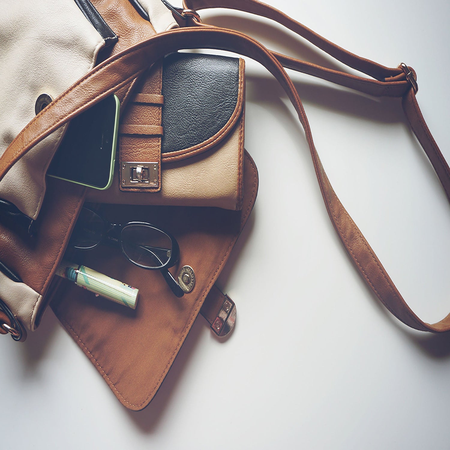 Brown leather handbag, wallet, glasses, phone on white table.