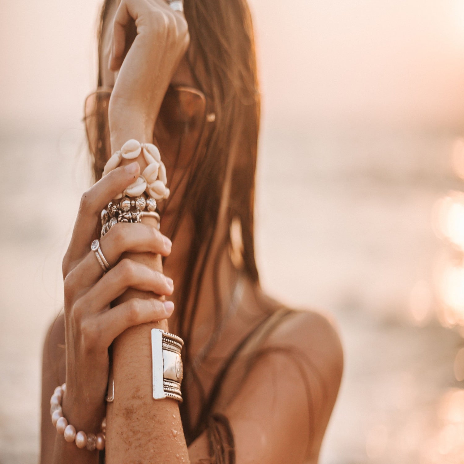 Woman holding wrist displaying bangles, bracelets, watch, earrings and necklace in sunset beach setting.