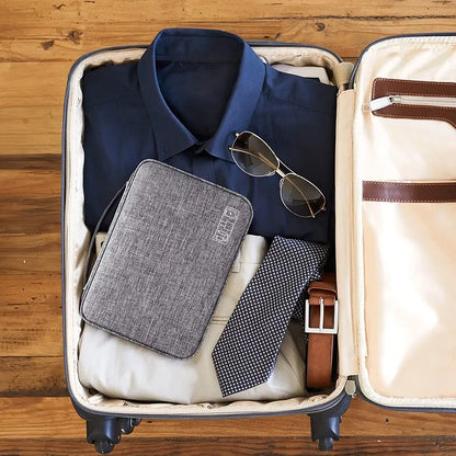 A packed suitcase featuring a blue shirt, sunglasses, a tie, trousers, a leather belt, and a gray tech organizer.