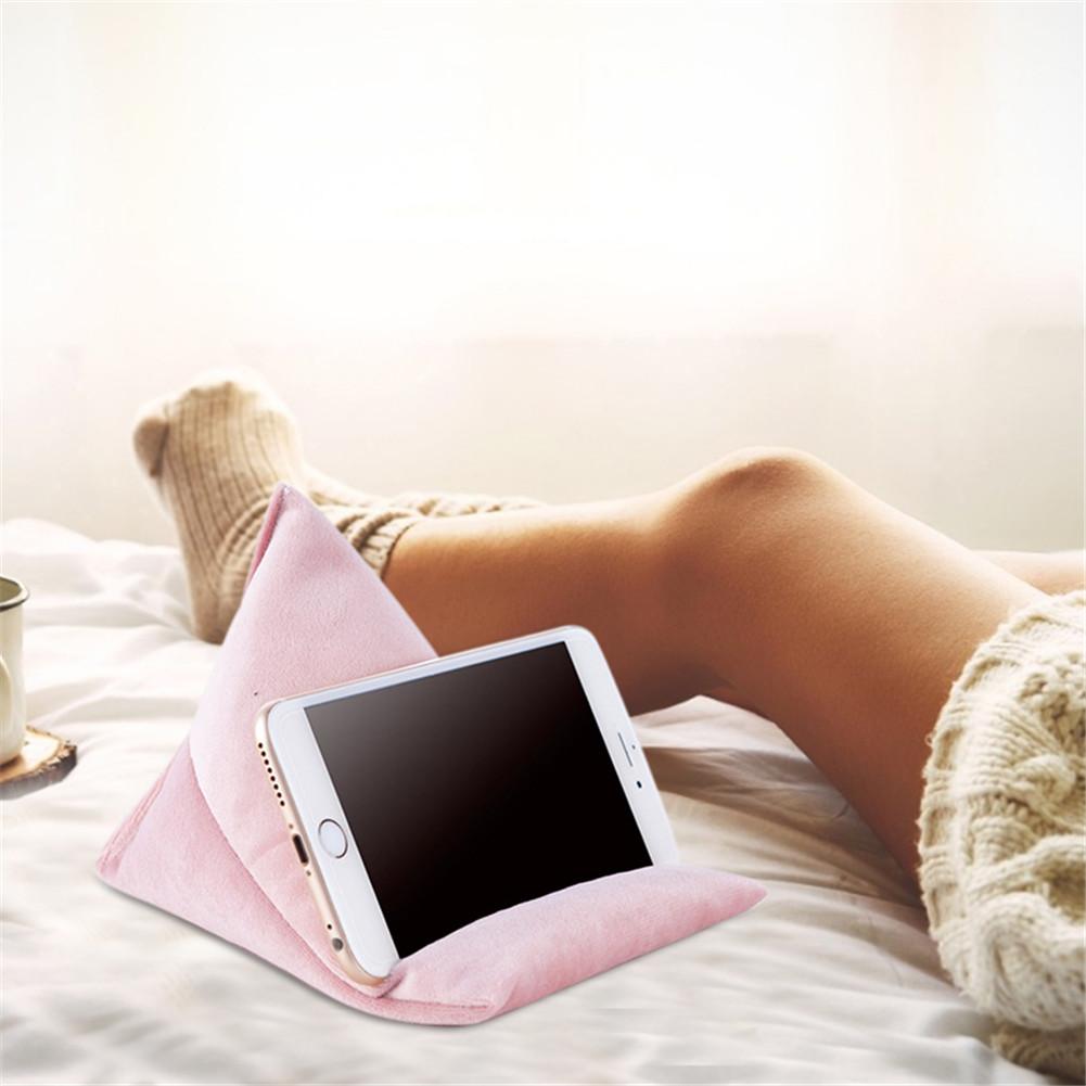 A pink phone stand rests on a bed, holding a smartphone. A person in cozy socks relaxes nearby, with a coffee cup in the background.