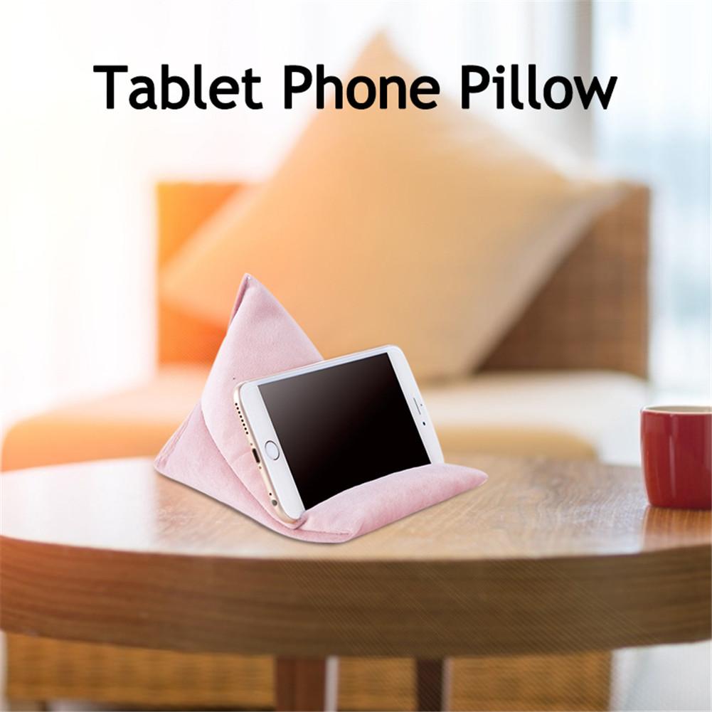 A pink tablet phone pillow propping up a smartphone on a wooden table, with a cozy sofa and mug in the background.