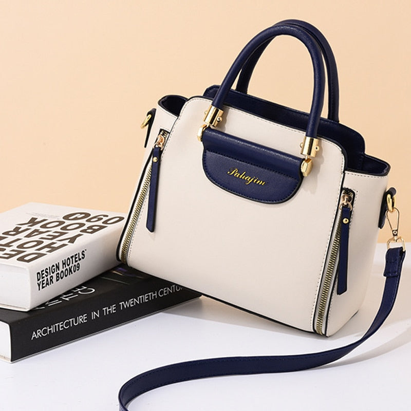 A stylish beige handbag with navy accents and gold detailing, displayed on a stack of fashion books against a soft peach background.