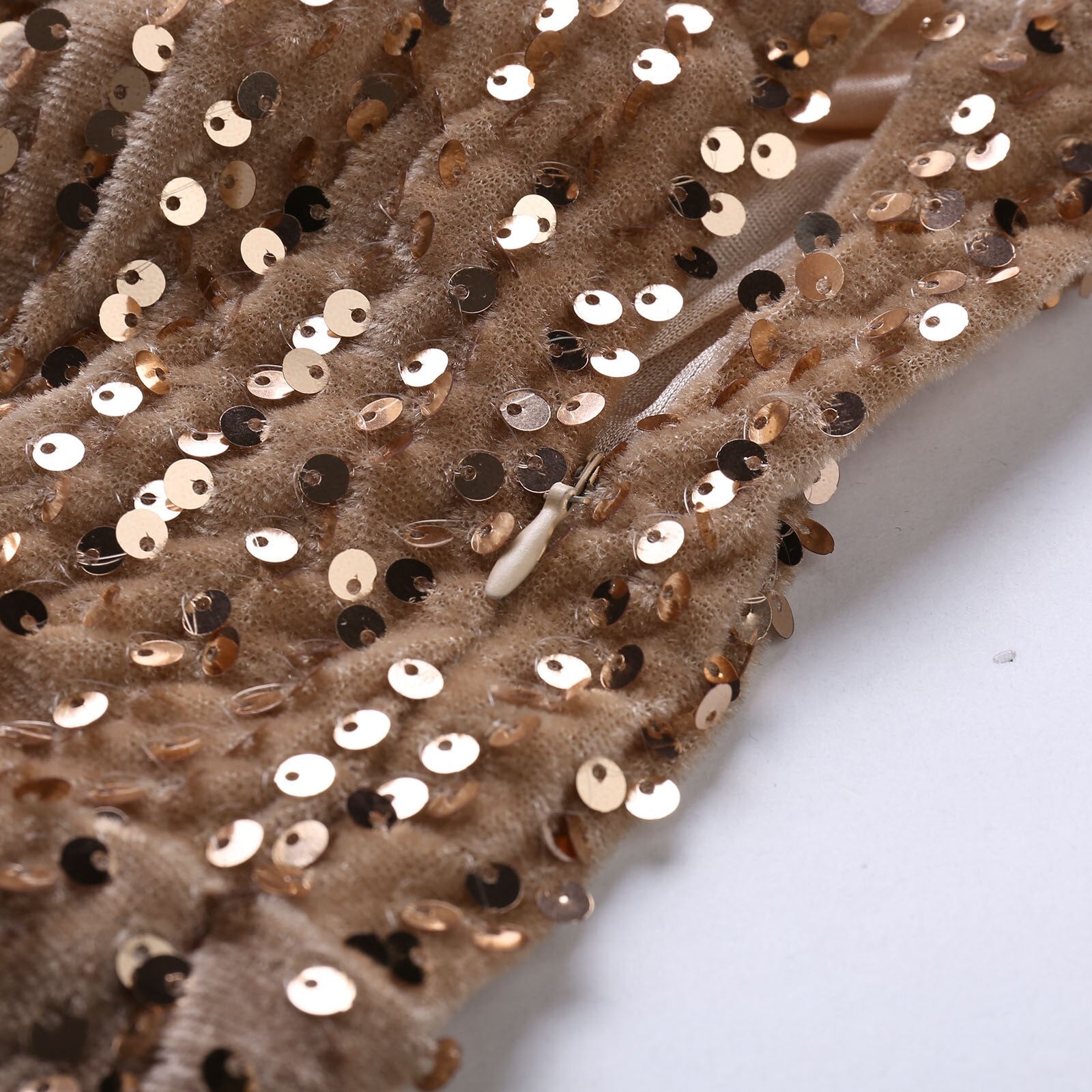 Close-up of a beige fabric adorned with shiny gold sequins, creating a textured and elegant appearance. A small zipper is partially visible.
