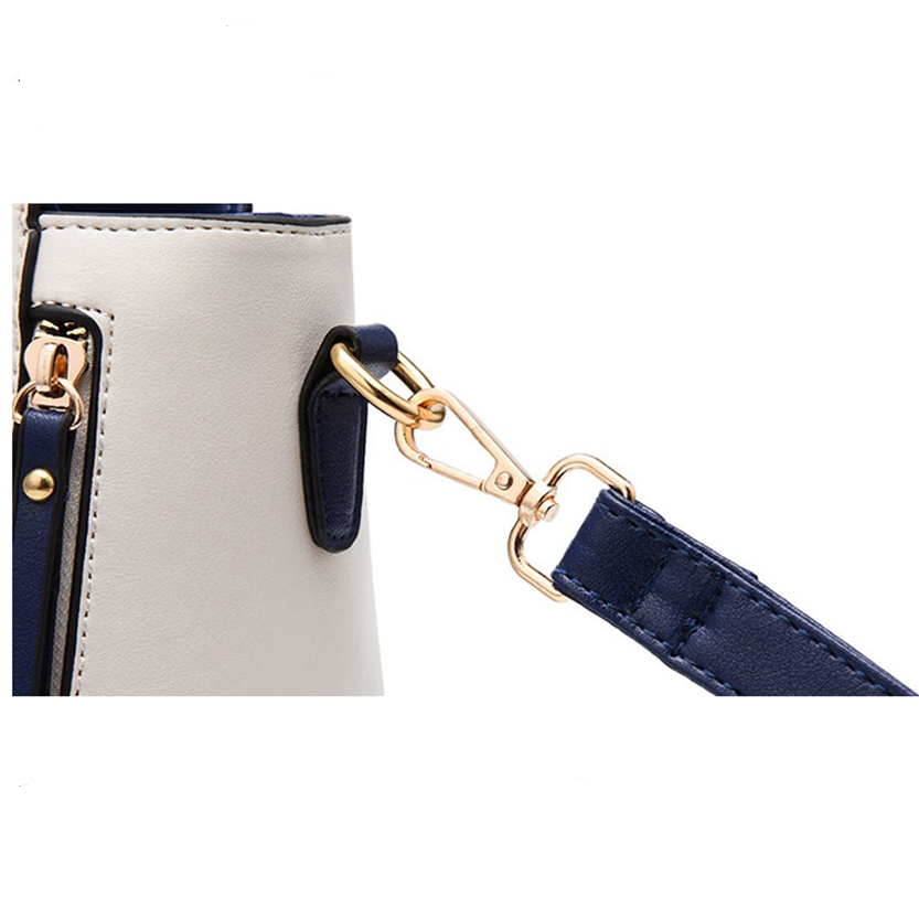 Close-up of a cream-colored handbag with a gold clasp attaching a navy blue strap to a metal ring. A zipper and dark blue trim are also visible.