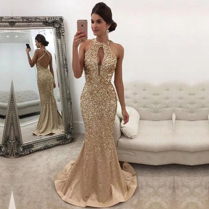 A woman wearing gold gown. The dress has a halter neck and fitted design, reflecting elegance. A plush sofa is in the background.