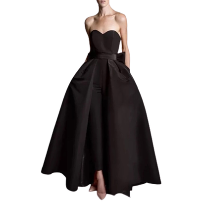 Strapless black gown with a fitted bodice and flared skirt, featuring a waist bow detail. Elegant and formal, paired with nude heels.