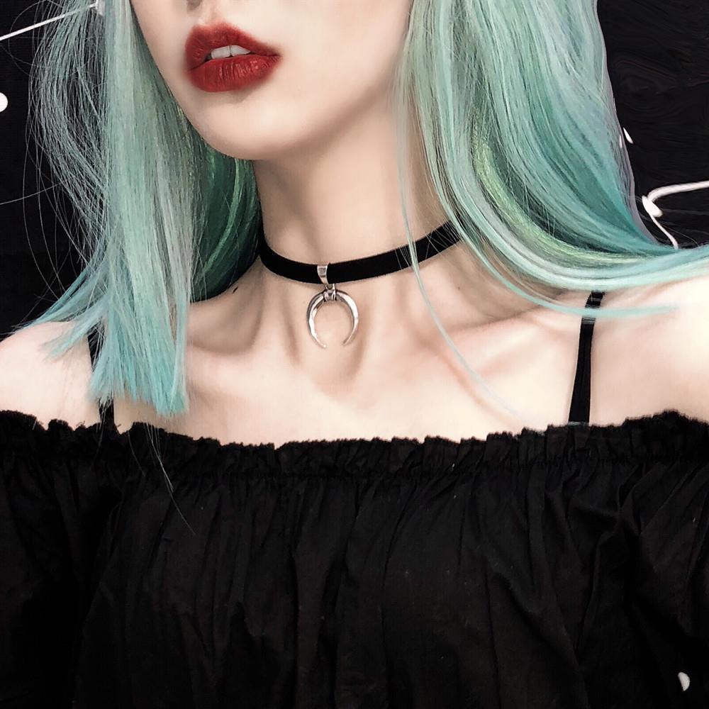 A woman with light blue hair wears a black off-the-shoulder top, red lipstick, and a black choker necklace featuring a silver crescent moon pendant.