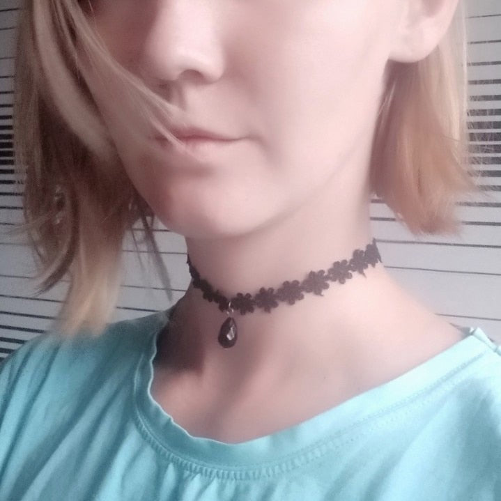 A woman wearing a black floral choker with a dark teardrop pendant, short blond hair, and a light blue shirt. The background features horizontal stripes.