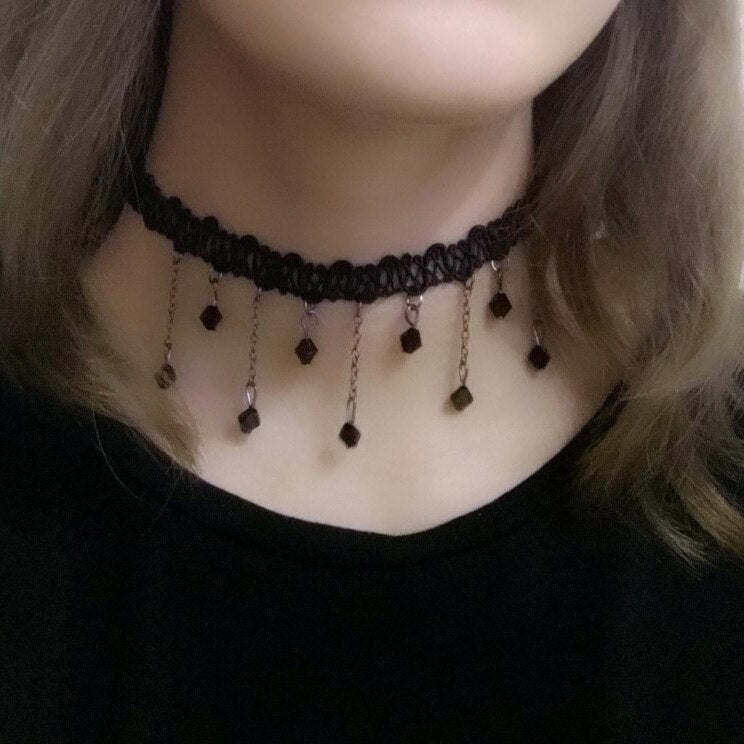 A woman wears a black lace choker with small dangling chain pieces, each ending in a black bead. The mood is elegant and gothic.
