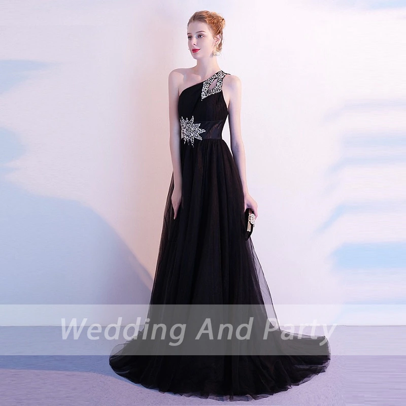 A woman wearing a black, one-shoulder evening gown with silver embellishments, holding a clutch. The backdrop is soft, conveying a formal tone.