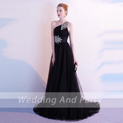A woman wearing a black, one-shoulder evening gown with silver embellishments, holding a clutch. The backdrop is soft, conveying a formal tone.