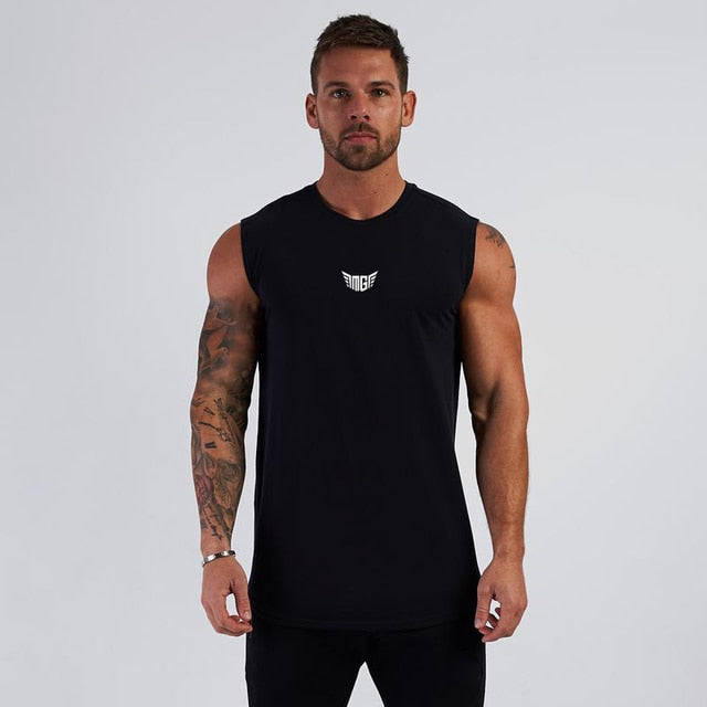 A muscular man stands confidently in a black sleeveless shirt with a small white logo, set against a plain gray background.