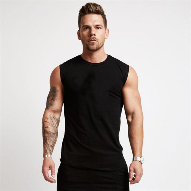 A muscular man in a fitted black sleeveless shirt stands confidently, showcasing his tattoos and a silver wristwatch.