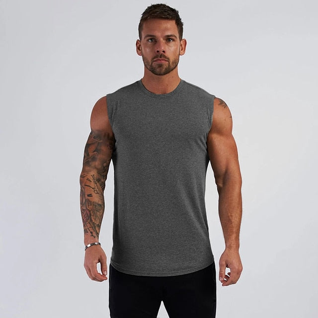 A muscular man stands confidently, wearing a gray sleeveless shirt, showcasing his tattoos against a plain background.