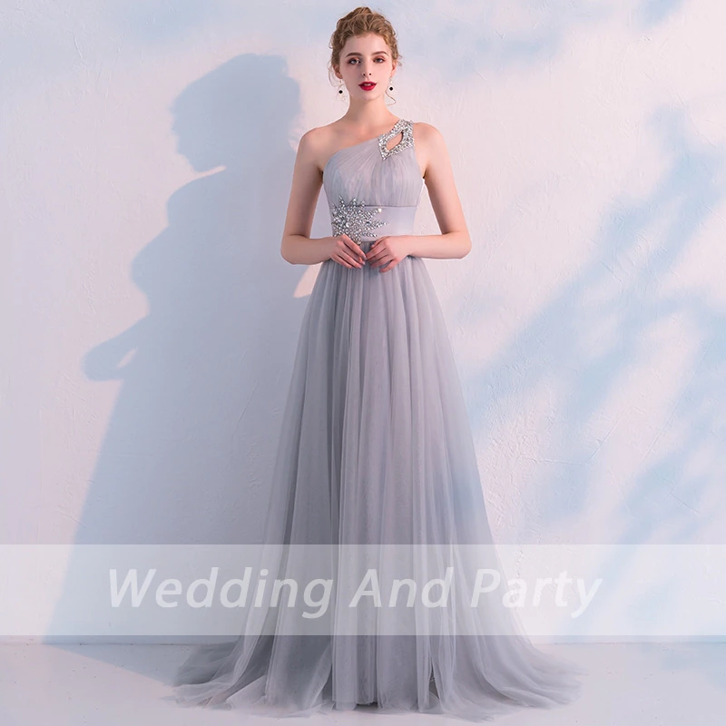 woman wearing a light blue, one-shoulder gown adorned with silver floral embroidery stands gracefully against a textured wall, exuding elegance.