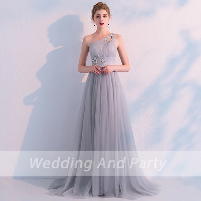 woman wearing a light blue, one-shoulder gown adorned with silver floral embroidery stands gracefully against a textured wall, exuding elegance.