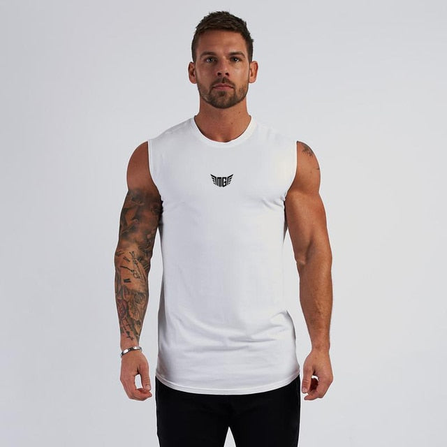 A muscular man stands in a sleeveless white shirt, showcasing his tattoos against a neutral backdrop.