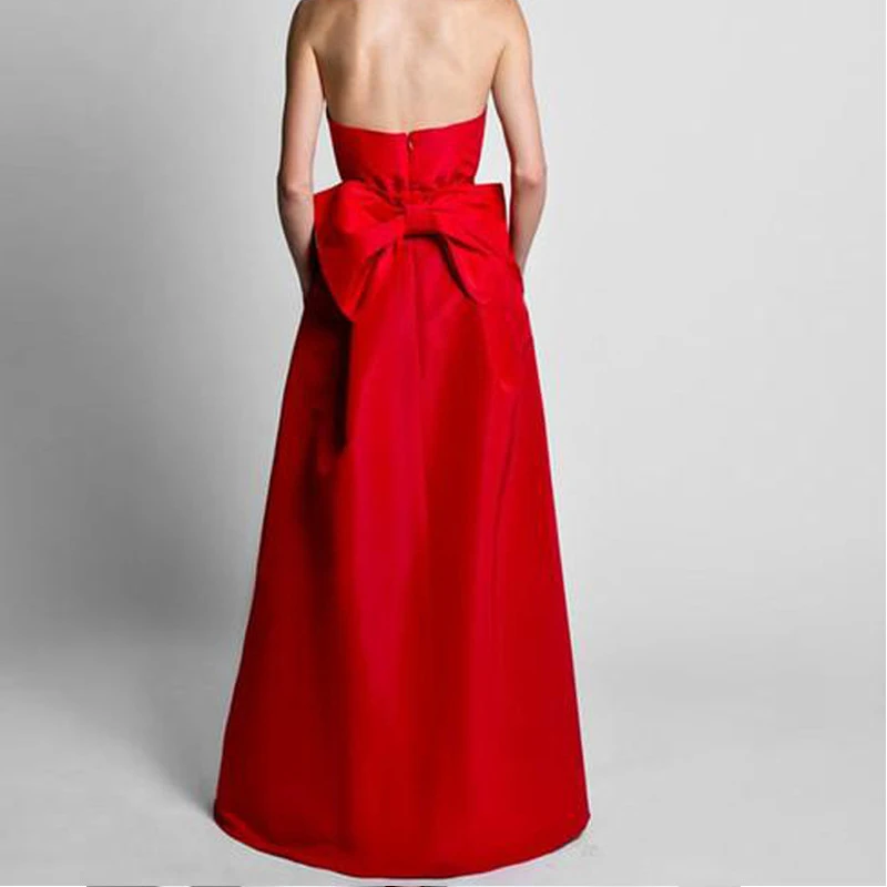 A woman in a strapless red gown with a large bow on the back stands against a plain gray background. The dress exudes elegance and sophistication.