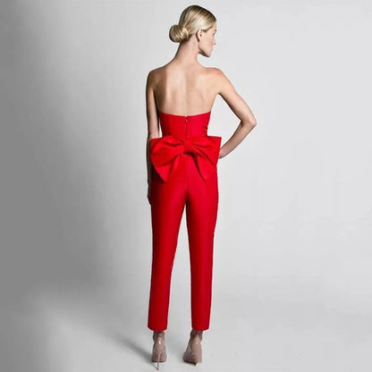 A woman in a strapless red jumpsuit with a large bow on the back stands against a plain background