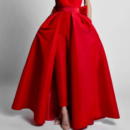 A woman wearing a striking red gown with a voluminous overskirt and tailored pants is standing against a plain gray background. Her posture is poised and elegant.