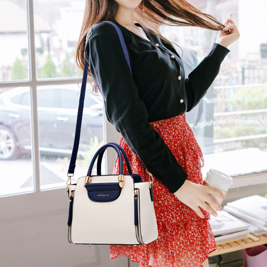 A woman in a black cardigan and floral skirt carries a stylish white and navy handbag and a coffee cup, standing by a window.
