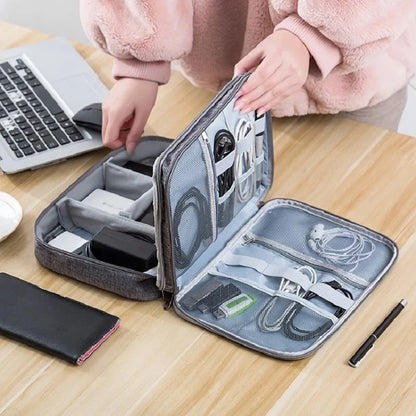 A person organizing tech accessories in a gray storage case, surrounded by a laptop, phone, and pen on a wooden table.