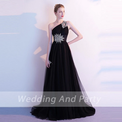 A woman in an  elegant black, one-shoulder evening gown with intricate silver embellishments stands against a light background, exuding sophistication.