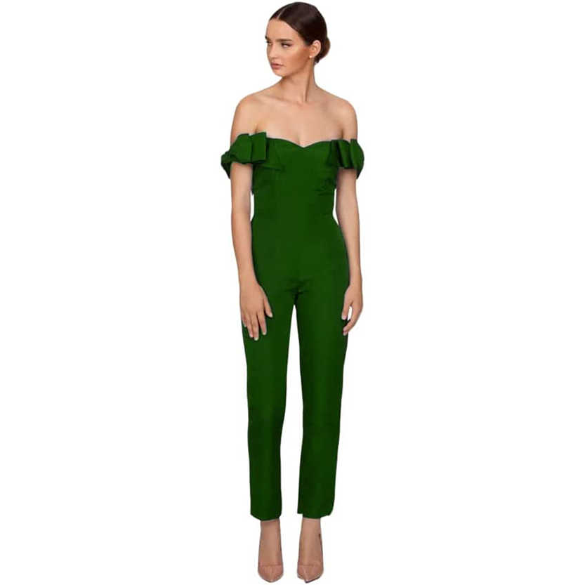 Woman wearing an off-shoulder green jumpsuit with ruffle details and neutral heels, standing against a white background. Elegant and poised.