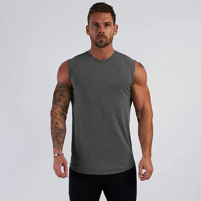 A muscular man stands confidently in a sleeveless gray shirt, showcasing intricate tattoos on his arms against a plain backdrop.