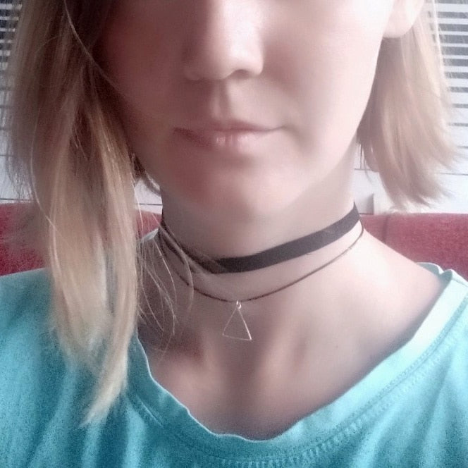 A woman with shoulder-length blonde hair wears a light blue shirt and two chokers. One choker is black, and the other features a small, silver triangle pendant. The background is softly blurred, conveying a calm atmosphere.