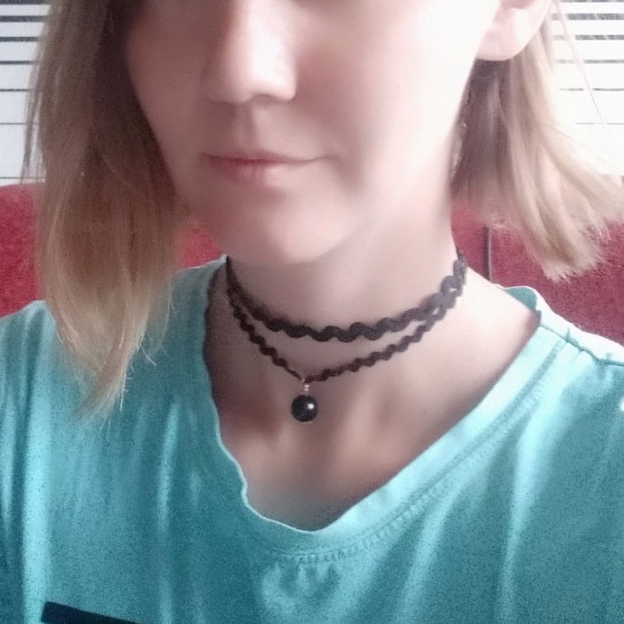 A woman with shoulder-length hair wears a turquoise shirt and A woman with shoulder-length hair wears a turquoise shirt and black choker necklaces with a pendant, set against a blurred red and striped background.with a pendant, set against a blurred red and striped background.