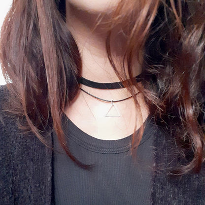 A woman wearing two necklaces: a black choker and a delicate chain with a  They have wavy, brown hair and wear a black top.