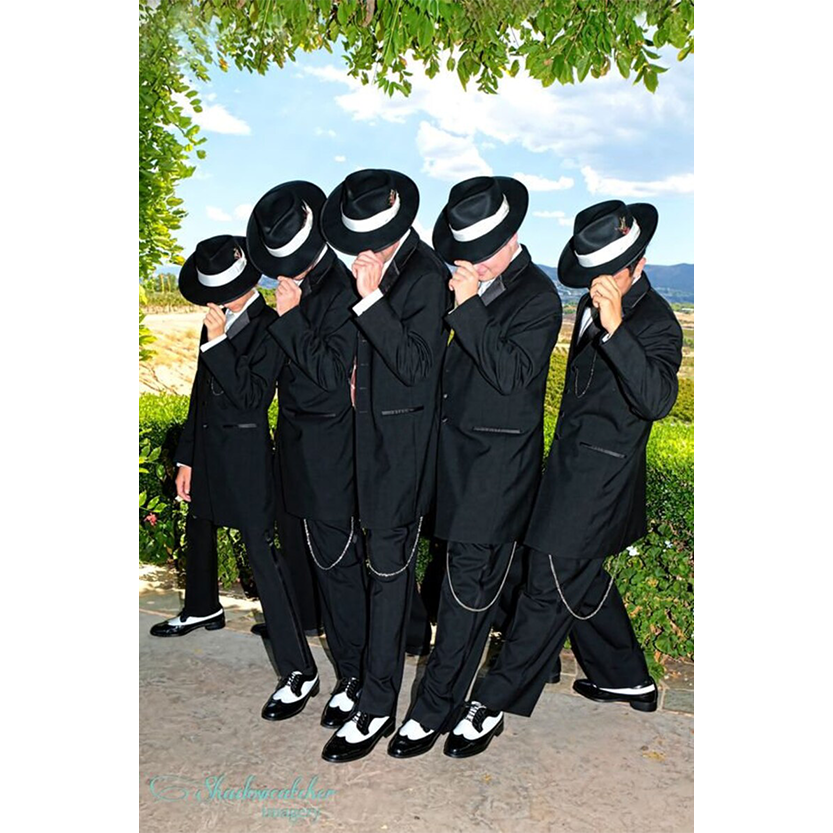 Five people dressed in black suits and white hats lean synchronously on a sunny day, set against a lush green backdrop under a leafy arch.