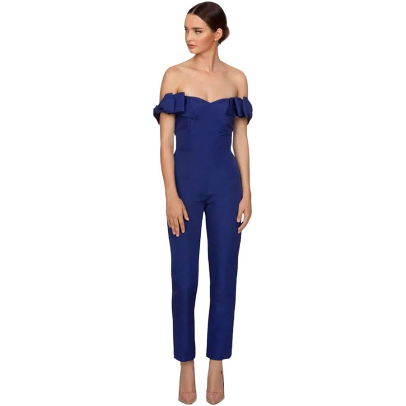 Woman in an off-shoulder, fitted blue jumpsuit with ruffled sleeves, stands with a neutral expression. She wears nude heels against a white background.
