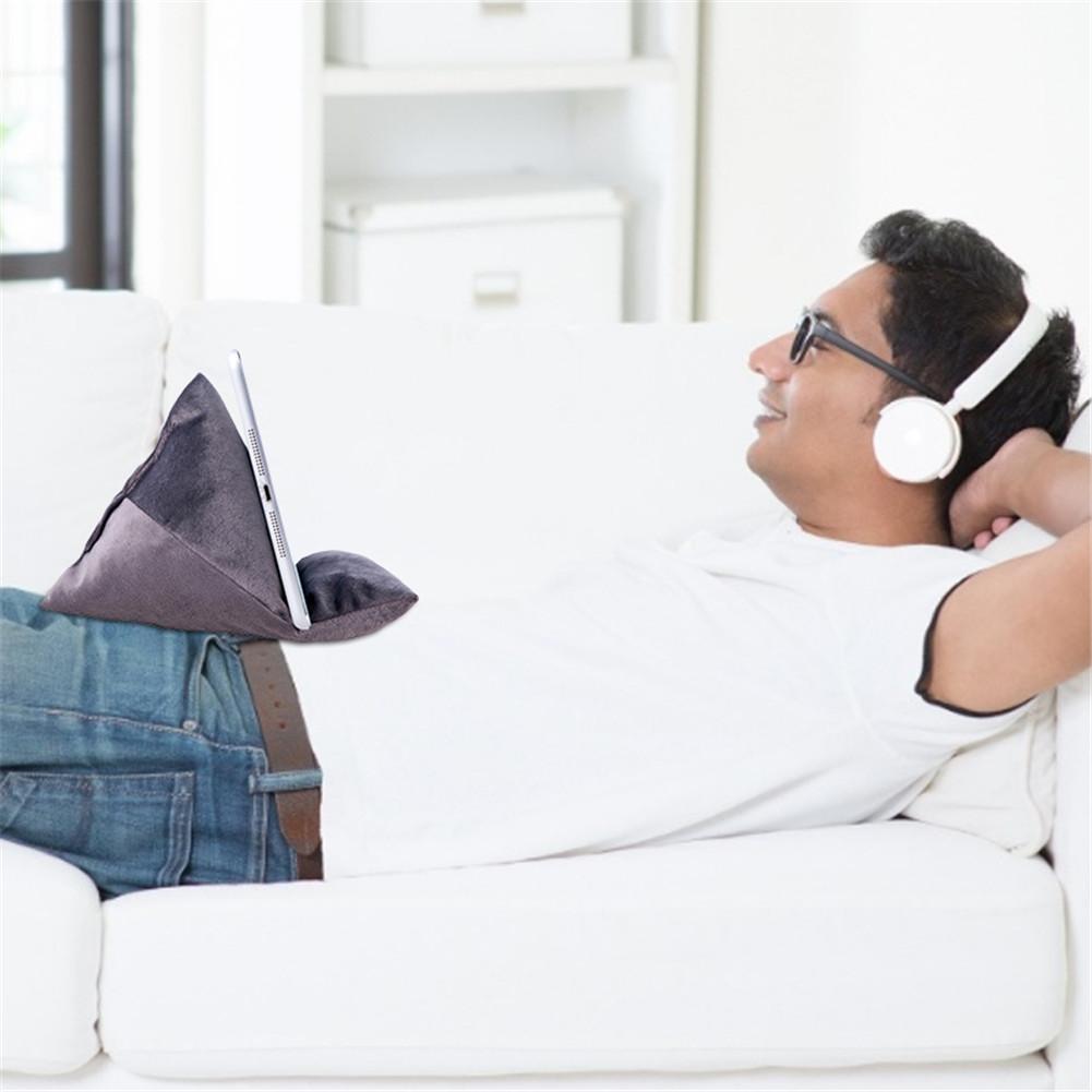 A person relaxes on a white couch, using a tablet propped on a triangular, soft cushion while wearing headphones.