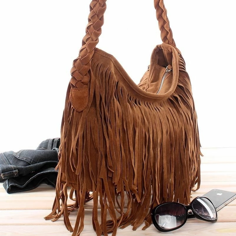 A stylish brown suede fringe handbag with braided handles, resting near sunglasses and denim on a light wooden surface.