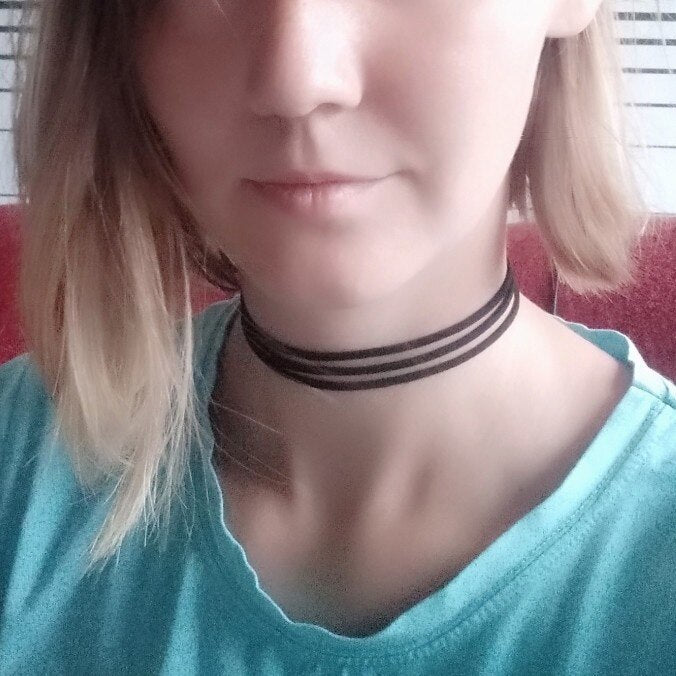 Woman wearing a blue shirt and a black choker necklace with multiple thin bands. Light brown hair partially visible, with a neutral expression.