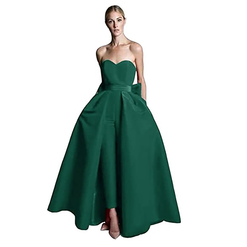 Woman in strapless emerald green gown with a fitted bodice and flared skirt, hands in pockets. Elegant and sophisticated tone, against a white background.