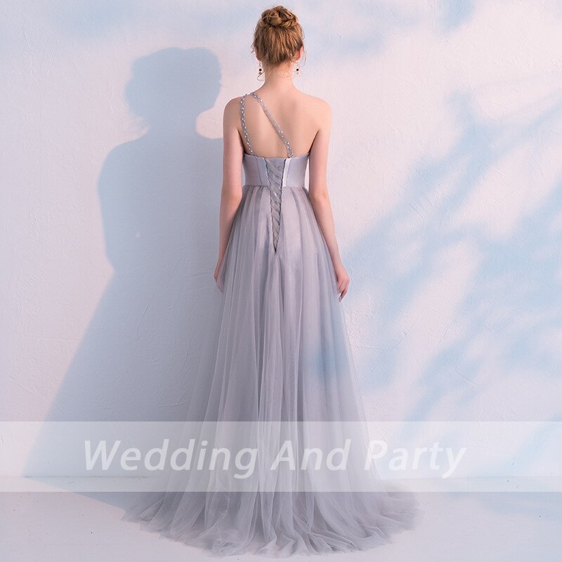 Woman in elegant, floor-length gray gown with beaded straps stands against a light wall, her hair in an updo, creating a graceful silhouette.