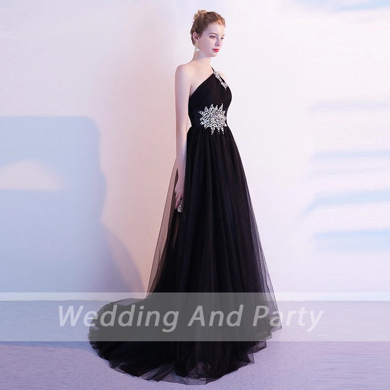 A woman wearing, floor-length, one-shoulder black gown with sparkling embellishments stands against a soft light background, exuding sophistication.