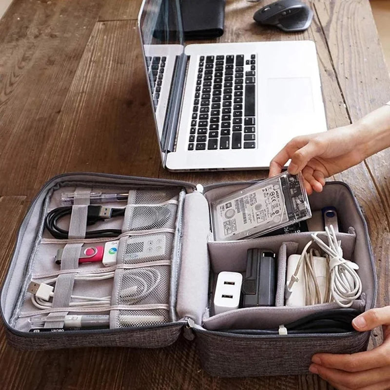 A person organizing electronic cables and devices in a gray travel case beside a laptop on a wooden table.