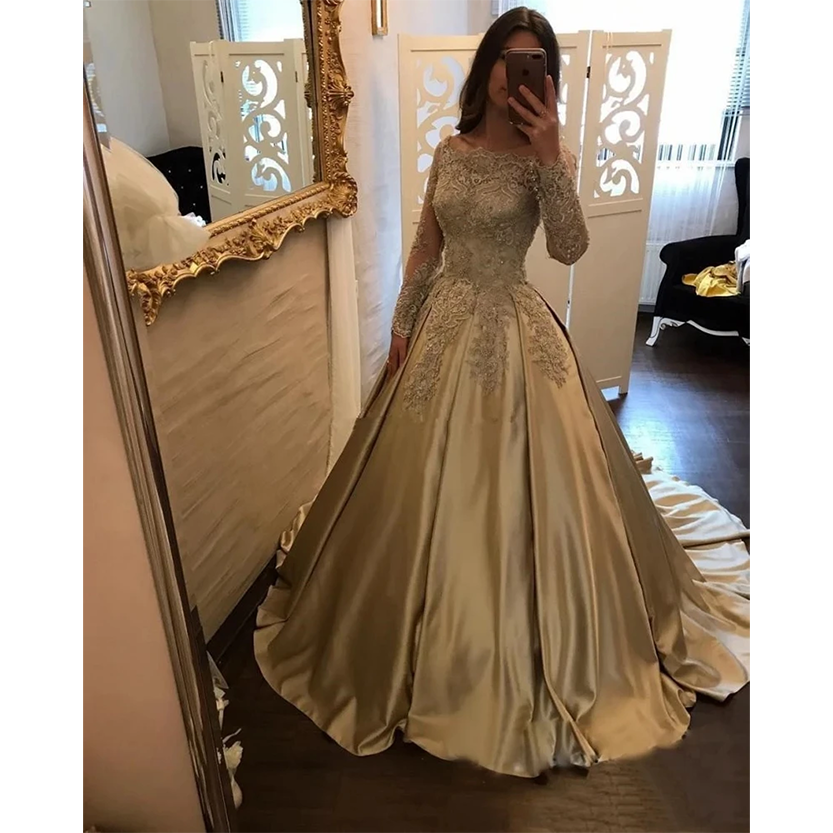 A woman stands in front of a mirror, wearing an elegant, gold lace gown with a full skirt and long sleeves. The room is softly lit, creating a luxurious, sophisticated ambiance.