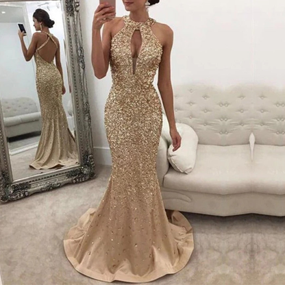 A woman wearing gold sequined evening gown n stands in a room with white walls.She poses elegantly beside a white tufted sofa.