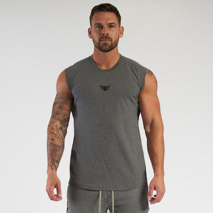 A muscular individual stands confidently wearing a sleeveless gray shirt and light gray shorts, showcasing his tattoos.
