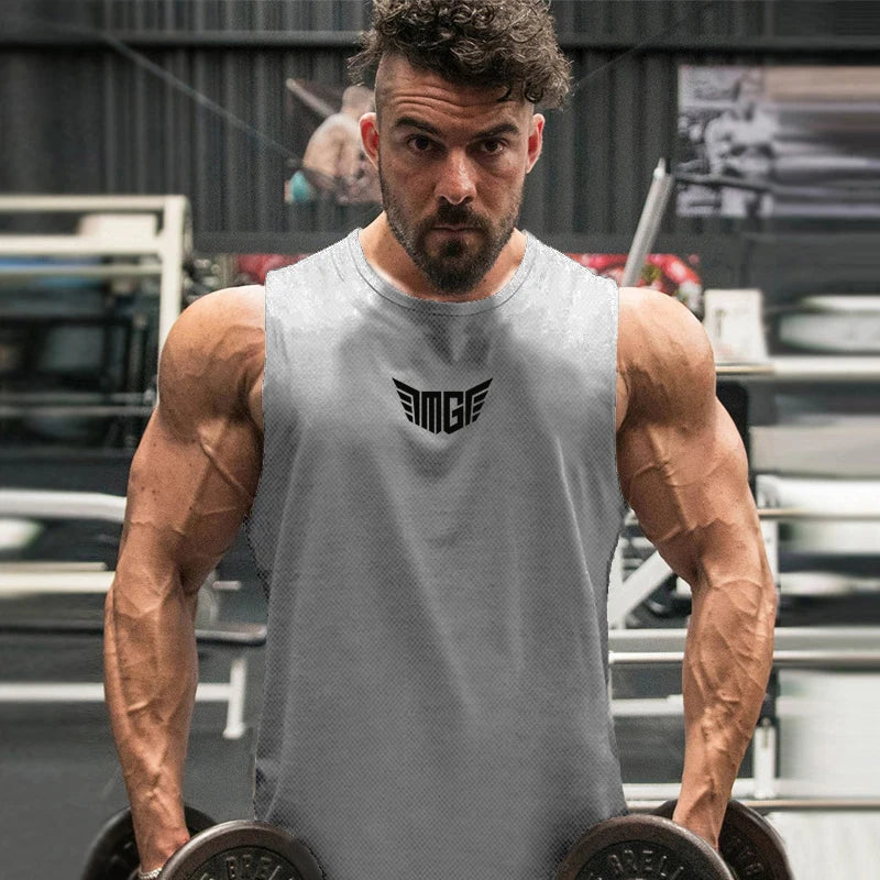 A muscular individual holds dumbbells, showcasing defined arms, wearing a gray sleeveless shirt with a logo. Gym setting in the background.