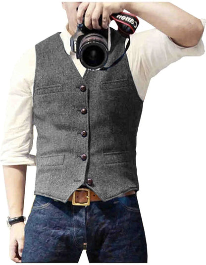 A man with a camera poses, wearing a gray vest over a white shirt, paired with blue jeans and a brown belt.