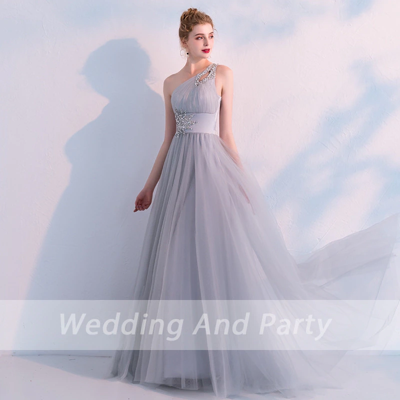 Elegant woman in a flowing, one-shoulder, light gray evening gown with jeweled details. She stands gracefully against a soft, pastel backdrop. 