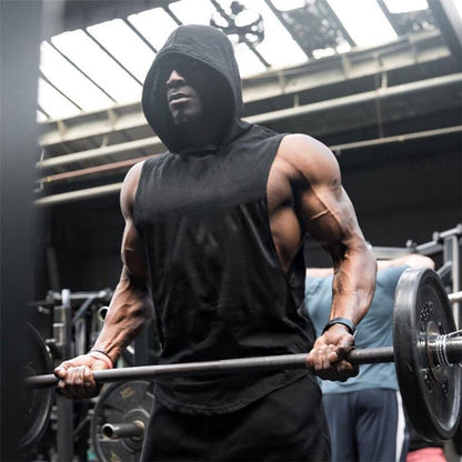 A muscular individual in a sleeveless hoodie lifts a barbell in a gym, showcasing strength and focus amidst training equipment.