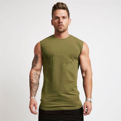 A muscular man poses confidently in a sleeveless olive green shirt and black shorts, showcasing his tattoos and stylish watch.