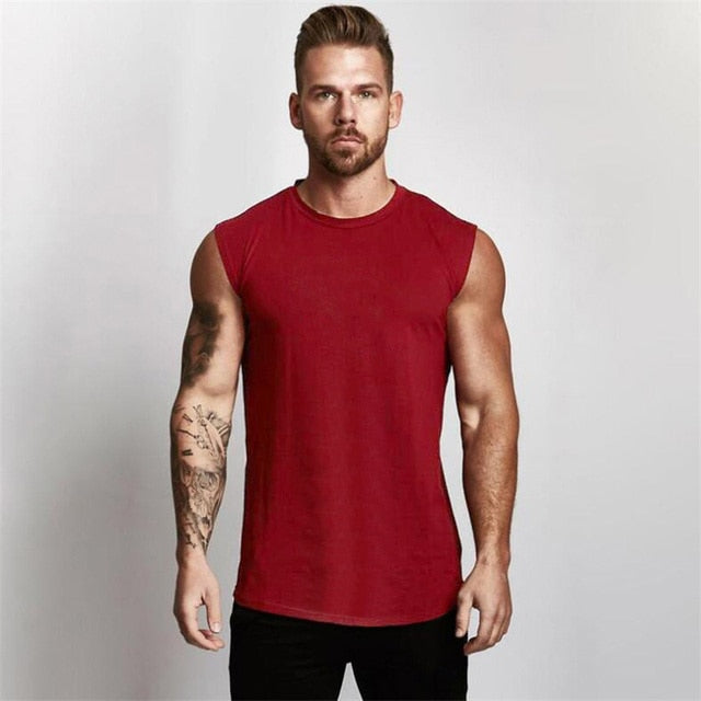 A muscular individual wearing a sleeveless red shirt stands confidently against a plain background, showcasing detailed arm tattoos.